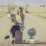 An image of a girl reading a map sitting on her suitcase in the middle of the road