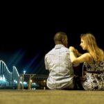 A woman sitting next to a man while putting her hands on his right shoulder as looking at him