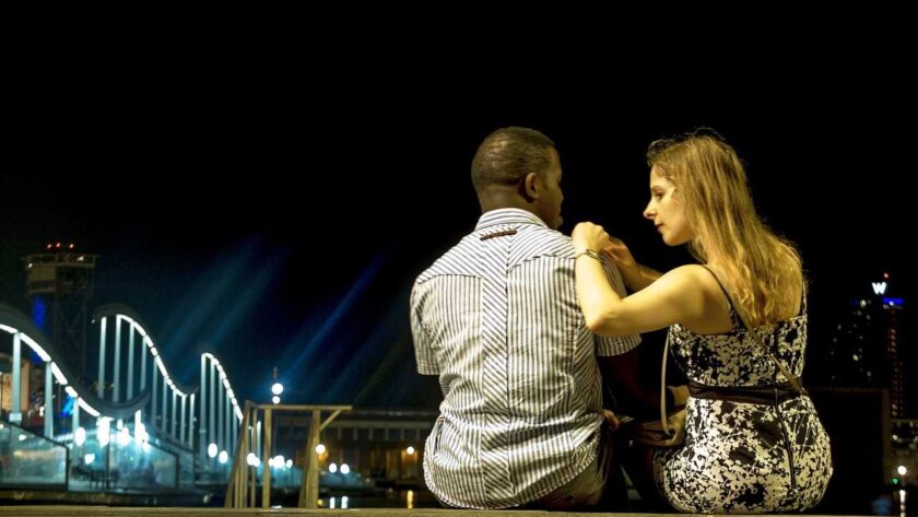 A woman sitting next to a man while putting her hands on his right shoulder as looking at him