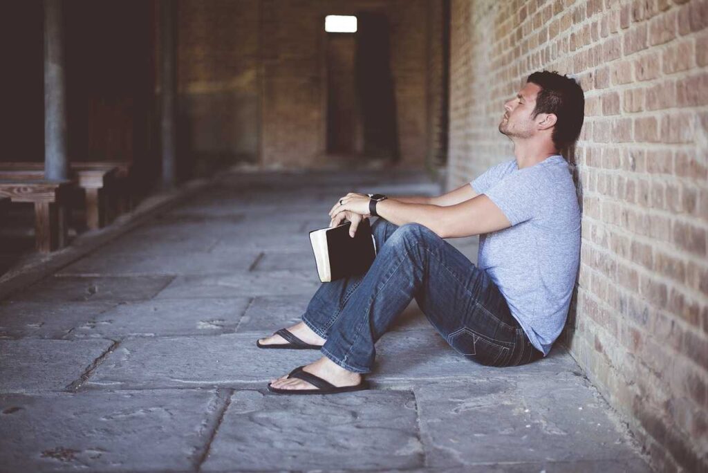 A man sitting on the ground leaning on a wall while holding a book 