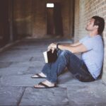 A man sitting on the ground leaning on a wall while holding a book