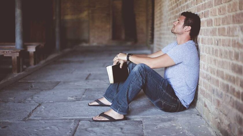 A man sitting on the ground leaning on a wall while holding a book