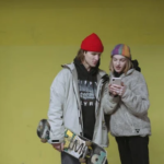 a woman standing next to a man holding a skate board, she is showing him something on a phone