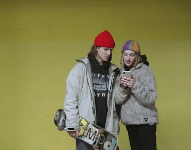 a woman standing next to a man holding a skate board, she is showing him something on a phone